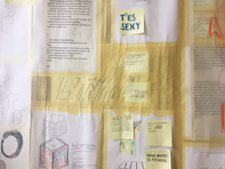 An einer Wand ist eine Collage aus handschriftlichen und gedruckten Notizen, Zeichnungen und Skizzen angebracht, darunter auch Haftnotizen mit Sätzen in verschiedenen Sprachen.