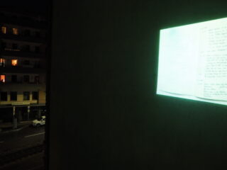 Ein schwach beleuchteter Raum mit einer Projektion eines handgeschriebenen Textes an einer Wand; eine nächtliche Stadtstraße mit geparkten Autos und Wohnhäusern, die durch ein Fenster sichtbar sind.