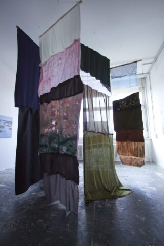 In einem Kunstatelier mit Betonboden und weißen Wänden hängen mehrere große Stoffbahnen in unterschiedlichen Farben und Texturen vertikal von der Decke.