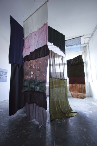 In einem Kunstgalerieraum mit weißen Wänden und Betonboden hängen Stoffpaneele in verschiedenen Farben und Texturen vertikal von der Decke.