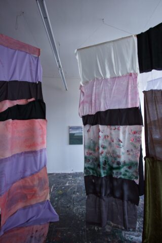 In einem Kunstatelier hängen Stoffbahnen in unterschiedlichen Farben und Mustern von der Decke, im Hintergrund ist an der Wand ein kleines Gemälde zu sehen.