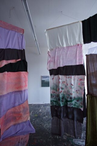 In einem Kunstatelier hängen bunte Stoffbahnen aus mehreren Lagen. Im Hintergrund ist ein kleines Gemälde an einer weißen Wand zu sehen. Der Boden ist mit Farbe bespritzt.