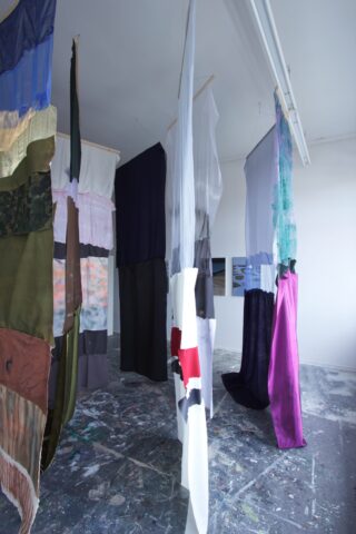 In einem Kunstatelier mit farbbespritztem Boden und weißen Wänden hängen lange Stoffbahnen in verschiedenen Farben und Texturen von der Decke.