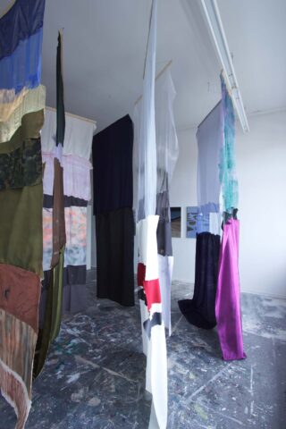 In einem Kunstatelier mit farbbespritztem Boden hängen Stoffbahnen in verschiedenen Farben und Mustern vertikal von der Decke.