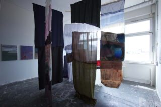 Mehrere große, halbtransparente Stoffbahnen hängen von der Decke eines hellen Raums mit weißen Wänden, sichtbaren Farbflecken auf dem Boden und Kunstwerken an den Wänden.