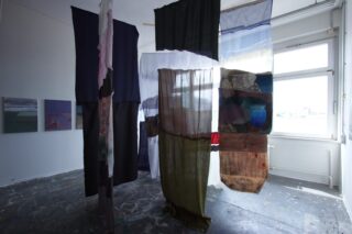 Mehrere große, halbtransparente Stoffbahnen hängen vertikal in einem Kunstatelier mit gemalten Kunstwerken an der Wand und natürlichem Licht, das durch ein Fenster einfällt.