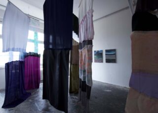 In einem Kunstatelier hängen Stoffpaneele in verschiedenen Farben von der Decke. An der Wand hängen zwei Landschaftsgemälde, und ein Betonboden ist zu sehen.