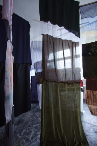 In einem Raum hängen verschiedene halbtransparente und undurchsichtige Stoffbahnen von der Decke, im Hintergrund fällt natürliches Licht durch ein Fenster.