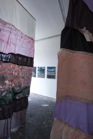 Im Vordergrund hängen zwei große Patchwork-Stoffbahnen, im Hintergrund sind an der weißen Wand eines Kunstateliers drei Landschaftsgemälde zu sehen.