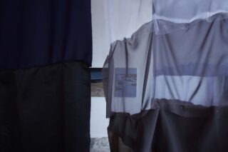 Vor einer kleinen, gerahmten Fotografie an einer weißen Wand hängen dunkle, durchscheinende Stoffbahnen.