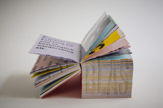 Ein offenes, buntes Buch mit dicken, ungleichmäßig geschnittenen Seiten, auf denen verschiedene Texte und Grafiken zu sehen sind, von der Seite betrachtet vor einem schlichten Hintergrund.