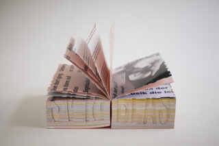 Ein Buch mit dicken, ungleichmäßig geschnittenen Seiten ist in der Mitte geöffnet und zeigt Text und ein auf die Seiten gedrucktes Schwarzweißfoto.