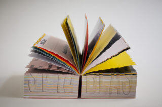 Ein offenes Buch mit Seiten in verschiedenen Farben und Texturen, das aufrecht steht und in dessen Ränder der Text „STAY DEAD“ eingeritzt ist.