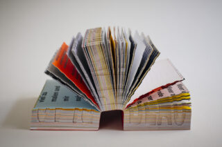 Ein dickes, mehrfarbiges, spiralgebundenes Buch steht offen auf seinem Rücken, mit ausgeschnittenem Text auf den Seiten und sichtbaren gedruckten Wörtern in verschiedenen Farben.
