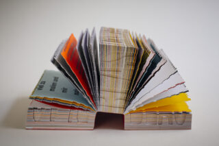 Ein dickes, buntes Buch steht aufrecht mit aufgefächerten Seiten und zeigt an den Rändern und Oberflächen verschiedene Farben, Muster und Texte.