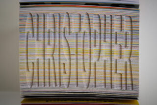 Ein Stapel farbiger Papierbögen, durch die in großen, vertikalen Buchstaben der Name „CHRISTOPHER“ geschnitten ist.