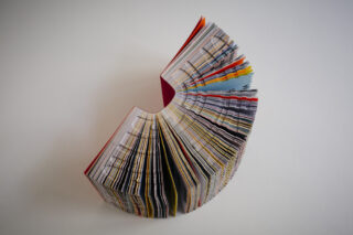 Ein dickes, buntes Buch steht aufrecht und offen, seine Seiten sind halbkreisförmig auf einem schlichten weißen Hintergrund aufgefächert.