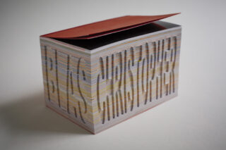 Ein dicker Stapel bunter Papierbögen bildet einen rechteckigen Block mit aufgedrucktem, vertikal verzerrtem Text an den Seiten und einem leicht angehobenen roten Blatt oben drauf.