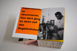 Ein aufgeschlagenes Buch zeigt eine Seite mit deutschem Text auf orangefarbenem Hintergrund und ein Schwarzweißfoto eines Mannes, der an einem Tisch mit verschiedenen Gegenständen arbeitet.