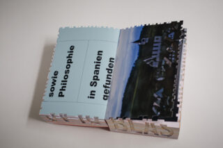 Offenes Buch mit hellblauem und schwarzem Text in deutscher Sprache auf der linken Seite und einem unscharfen Foto einer ländlichen Landschaft mit Gebäuden auf der rechten Seite.