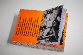 Ein Buch mit orangefarbenen, puzzleteilförmigen Seiten, auf denen auf einer Doppelseite eine aufgedruckte deutsche Nachricht und ein Schwarzweißfoto zu sehen sind.
