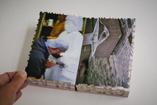 Eine Hand hält ein Buch mit zwei Fotos aufgeschlagen: eines zeigt eine Person, die eine weiße Statue formt, und ein anderes eine Steinbank auf einem gepflasterten Gehweg.