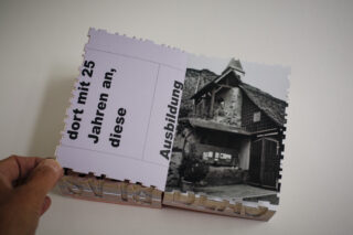 Eine Hand hält ein aufgeschlagenes Heft mit deutschem Text auf der linken Seite und einem Schwarzweißfoto eines rustikalen Gebäudes auf der rechten Seite.