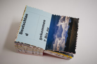 Ein Buch mit ausgeschnittenen, puzzleartigen Seiten ist geöffnet. Eine Seite enthält deutschen Text, eine andere zeigt das Foto eines bewölkten Himmels über einer Landschaft.