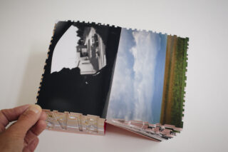 Eine Hand hält ein Fotobuch aufgeschlagen, in dem zwei Bilder zu sehen sind: ein Schwarzweißfoto der Silhouette einer Person in einem Auto und eine farbige Landschaft mit Feldern und Wolken.