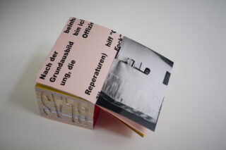 Ein kleines Heft mit deutschem Text auf dem Umschlag, das teilweise geöffnet ist und ein Schwarzweißfoto einer Person an einem Tisch im Inneren zeigt.