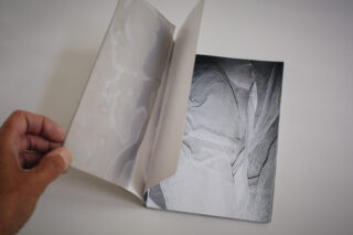 Eine Hand blättert eine Seite in einem Buch um und gibt den Blick auf ein abstraktes Graustufenbild mit netzartigen Texturen und geschichteten Stoffmustern frei.