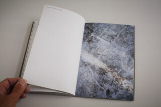 Eine Hand hält ein Buch aufgeschlagen, auf dem links eine leere Seite und rechts eine Seite mit einem strukturierten, abstrakten Bild in Grau und Weiß zu sehen ist.