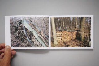 Eine Hand hält ein aufgeschlagenes Buch mit zwei Fotos von gestapelten Brennholzstapeln in einer Waldlandschaft im Freien, einer davon mit einer Plane abgedeckt, der andere freigelegt.