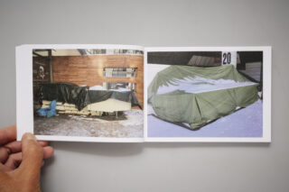 Eine Hand hält ein aufgeschlagenes Buch mit zwei Fotos von Objekten im Freien, die mit grünen Planen abgedeckt und von Schnee umgeben sind.