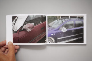 Eine Person hält ein aufgeschlagenes Buch mit zwei Fotos von Autos hoch, bei denen jeweils ein Seitenspiegel mit einer Plastiktüte abgedeckt ist.
