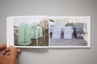 Eine Hand hält ein Buch aufgeschlagen, in dem zwei Fotos zu sehen sind: grüne zeltartige Strukturen auf der linken Seite und graue und violette kastenartige Unterstände auf der rechten Seite, beide im Freien.