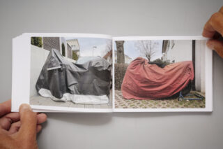 Zwei Hände halten ein Buch aufgeschlagen, in dem Fotos von mit Planen bedeckten Motorrädern zu sehen sind; eine Plane ist schwarz-weiß, die andere rot.