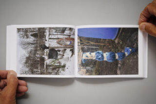 Eine Person hält ein Fotobuch aufgeschlagen, das zwei Außenaufnahmen zeigt: eine von einer schneebedeckten Szene mit Objekten und eine andere von einer vertikalen Anordnung aus blauem und weißem Stoff.