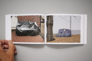 Ein offenes Buch mit zwei Fotos von mit Planen abgedeckten Objekten im Freien; eines befindet sich neben einer Ziegelmauer, das andere neben einem Baum und einer weißen Wand.