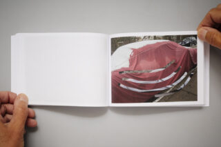 Eine Person hält ein aufgeschlagenes Buch mit dem Foto eines Autos auf einer Straße, das mit einer roten, zerrissenen Plane mit weißen Streifen bedeckt ist.
