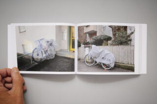 Eine Hand hält ein aufgeschlagenes Fotobuch mit zwei Bildern von Fahrrädern, die mit durchsichtigen Plastikplanen bedeckt und in der Nähe von Gebäuden geparkt sind.