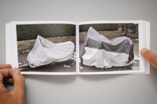 Eine Person hält ein aufgeschlagenes Buch mit einem Foto von zwei auf einer Straße geparkten Motorrädern hoch, die beide mit Schutzplanen abgedeckt sind.