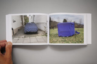 Eine Hand hält ein Buch aufgeschlagen, in dem zwei Fotos zu sehen sind: links ein mit einem blau gemusterten Tuch bedecktes Auto und rechts eine blaue zeltartige Struktur im Freien.