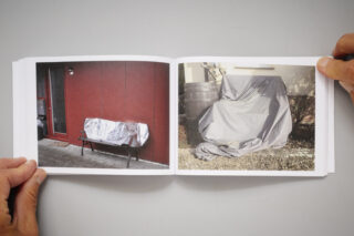 Eine Person hält ein aufgeschlagenes Buch mit Fotos von mit Planen abgedeckten Objekten in der Hand; ein Objekt liegt auf einer Bank, das andere neben einem Gebäude.