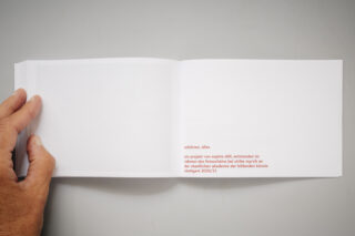 Eine Hand hält ein Buch aufgeschlagen, auf dem sich eine größtenteils leere Seite befindet. In der unteren rechten Ecke der rechten Seite ist ein kleiner orangefarbener Text auf Deutsch abgedruckt.