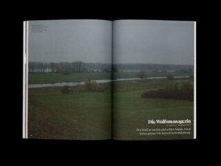 Eine Magazinseite zeigt eine wolkige Landschaft mit grünen Feldern, Bäumen und einem gewundenen Fluss. Der deutsche Text lautet „Die Wolfsmanagerin“ und darunter ein einführender Satz.