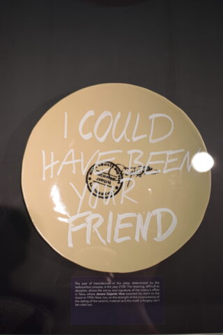 Auf einem beigefarbenen Teller in einer Museumsvitrine steht in der Mitte der in Weiß gemalte Schriftzug „ICH HÄTTE DEIN FREUND SEIN KÖNNEN“ über einem Stempel und einer handschriftlichen Notiz.