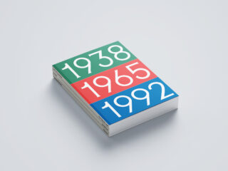 Ein Stapel Bücher, deren Einbände die Jahreszahlen 1938 in Grün, 1965 in Rot und 1992 in Blau zeigen, ordentlich angeordnet auf einem hellen Hintergrund.