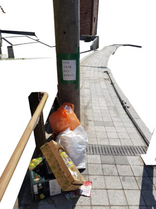 Ein Stapel Müllsäcke und -kartons steht am Fuß eines Pfostens mit einem grünen Schild auf einem gepflasterten Gehweg.