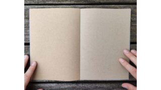 Zwei Hände halten ein leeres Notizbuch aus braunem Papier auf einer Holzoberfläche offen.